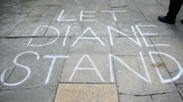 A grey pavement with the words "Let Diane Stand" written on it in chalk in large, block capital letters.