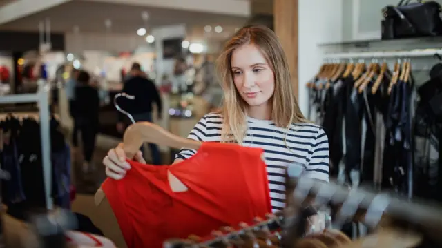 A woman standing in a clothes store with rails of clothing hanging around her. The woman is holding a clothes hanger with an orange garment on.