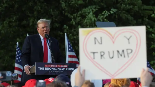 Donald Trump speaks at a campaign rally in The Bronx, New York; in the foreground, a supporter holds up a sign reading: "NY (heart) Trump"