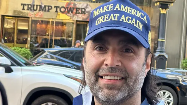 A man wearing a Make America Great Again hat poses outside of Trump Tower