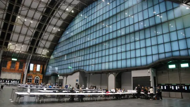 A big hall is seen with long tables where people are tabulating votes in Manchester