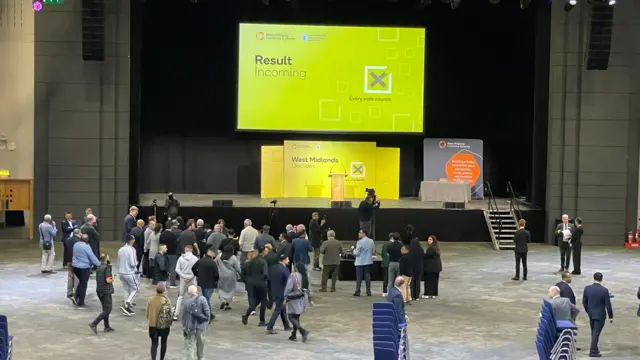 People begin gathering at the foot of a stage. Above the stage is a sign, reading: "Result incoming"