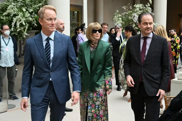 A white man in a blue suit, a white woman in sunglasses and a green coat with flowers on it and a white man in a black coat walk together