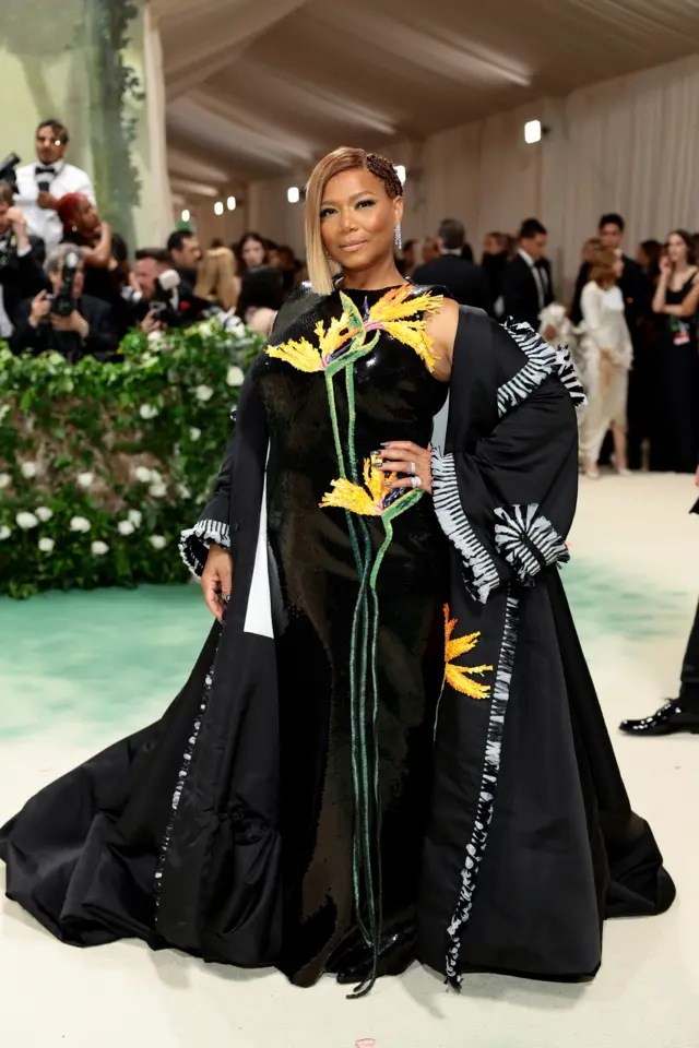 A Black woman wears a black beaded dress with large orange and yellow flowers on it