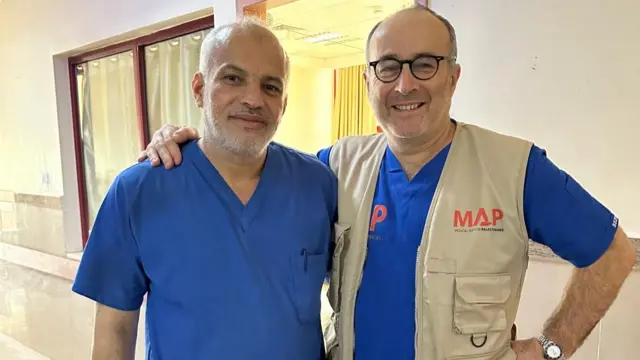 Two men in blue hospital scrubs smile at the camera