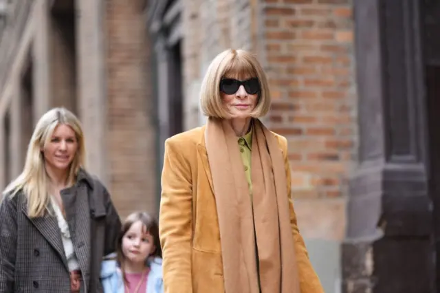 Anna Wintour wears sunglasses, a brown fringed long scarf, a brown / orange long fabric coat with shoulder pads, a khaki flowing skirt, beige snake print pattern pointed boots in leather, outside Proenza Schouler , during New York Fashion Week, on February 10, 2024 in New York City