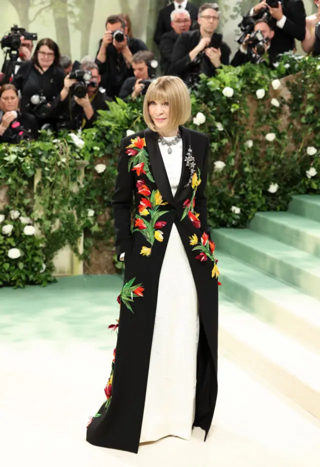 Anna Wintour stands in a white gown and black coat with floral appliques