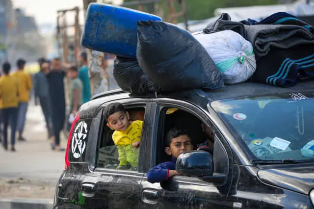 A boy peers out of the front seat of car, while another child pokes his head out of the backseat of the vehicle, which is carrying bags and blankets on its roof.