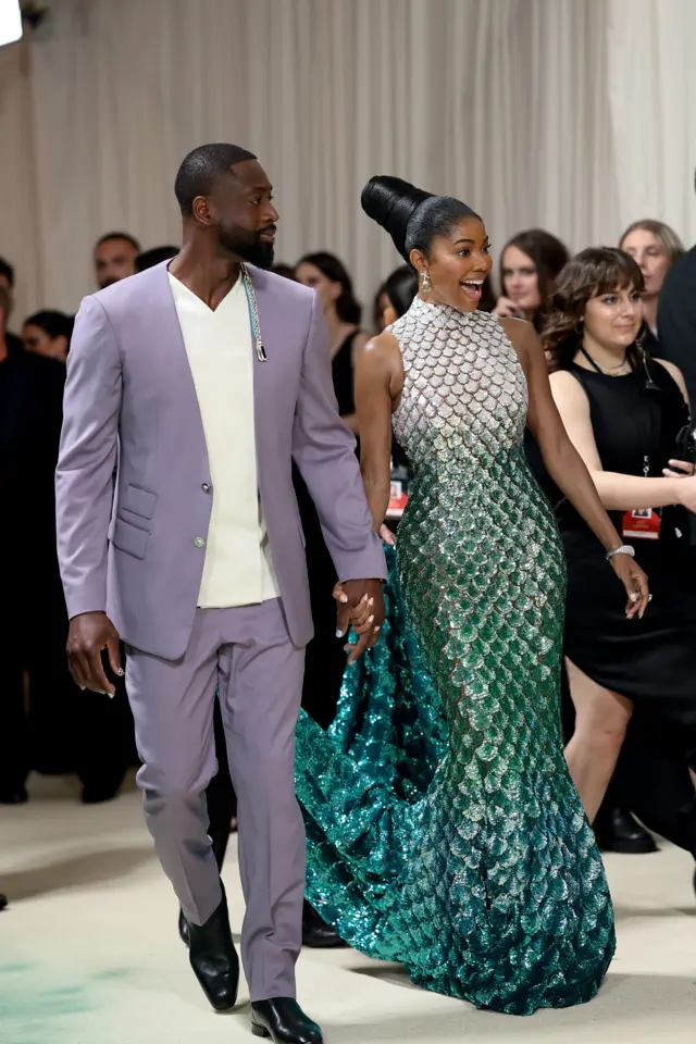 Dwyane  in a lavender suit walks with a black woman in a mermaid-like sequined gown in blue, white and teal