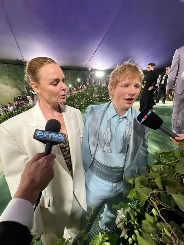 A white woman in a white suit stands with a white red-headed man in a powder blue suit