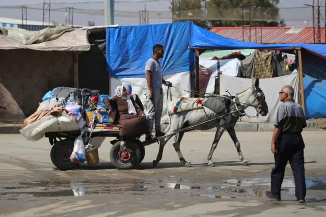 A man leads a donkey cart carrying two women and a pile of belongings. There are tents in the background.