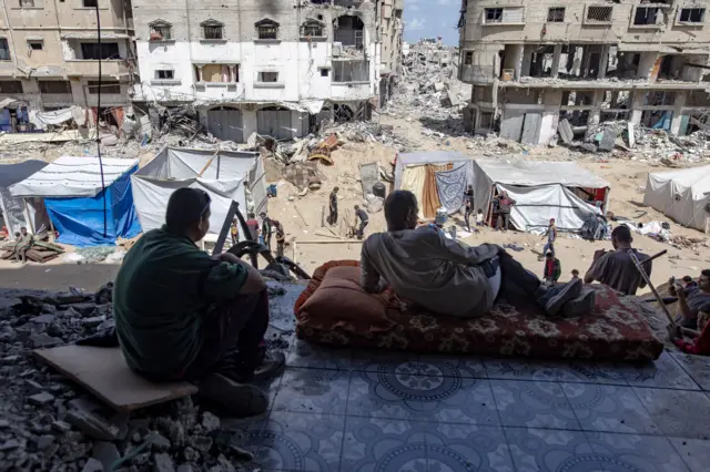 Two men overlooking the city of Khan Yunis, where people are setting up tents among ruins.