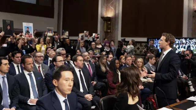 Mark Zuckerberg stands, speaking to a room of people, some of whom hold up photos of young people