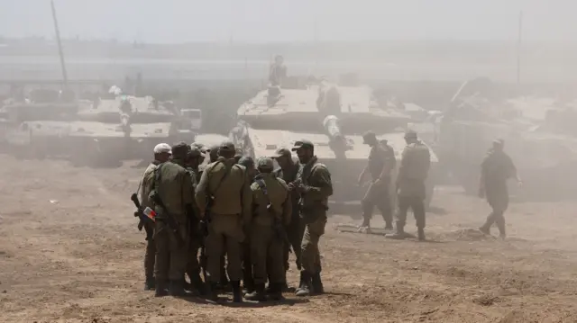 Several Israeli soldiers gather near the border fence with the Gaza Strip, with military vehicles in the background.