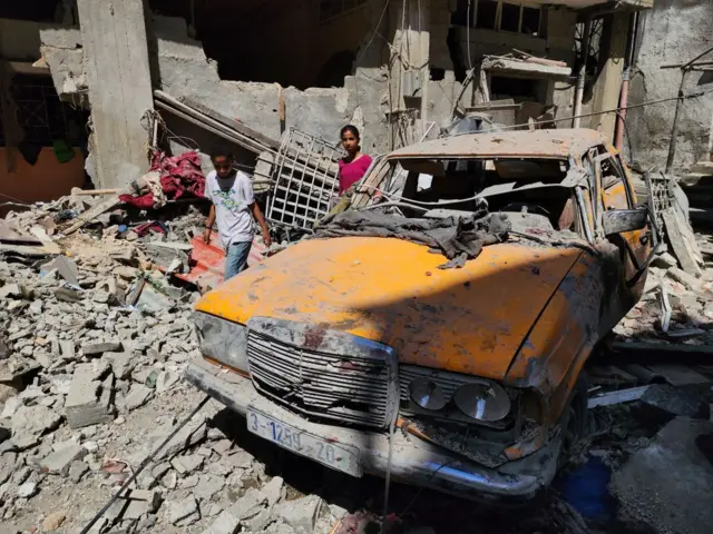 Children pick up some of their belongings from amid the rubble, and a destroyed yellow car is visible in the foreground