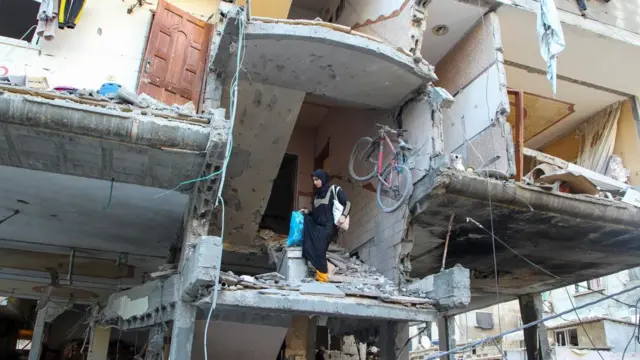 A Palestinian woman walks down the stairs of a house hit in an Israeli strike carrying bags of belongings.