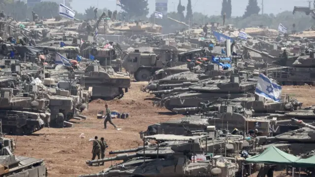 Israeli soldiers with military vehicles gather at an undisclosed position near the border fence with the Gaza Strip with Israeli flags positioned on top of some tanks.