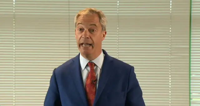 Nige Farage standing in front of some blinds