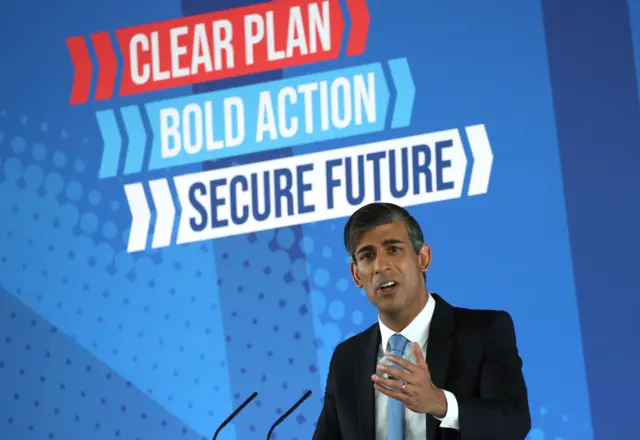 Sunak stands in front of a sign saying 'clear plan, bold action, secure future'