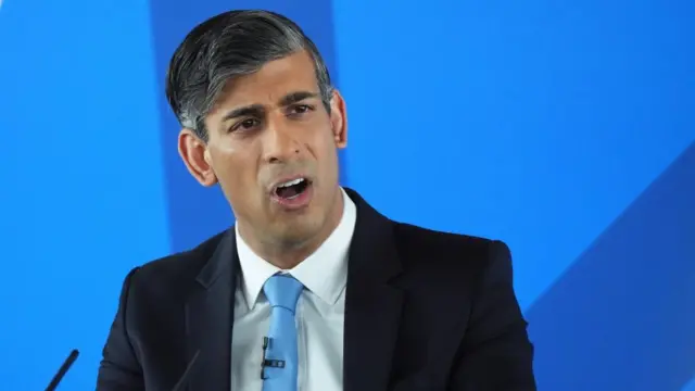 Rishi Sunak stands in front of an audience in a blue suit against a blue background