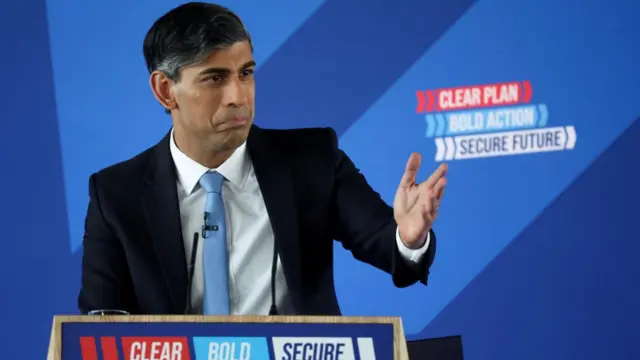 Rishi Sunak stands in front of a podium and gestures to the crowd.