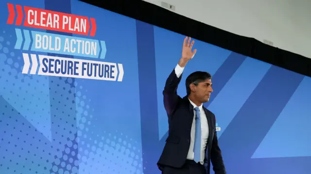 Sunak waves as he stands in front of a banner saying 'clear plan, bold action, secure future'