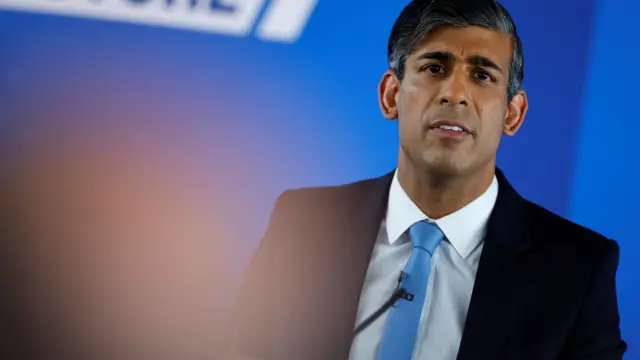 Rishi Sunak stands in front of a blue background