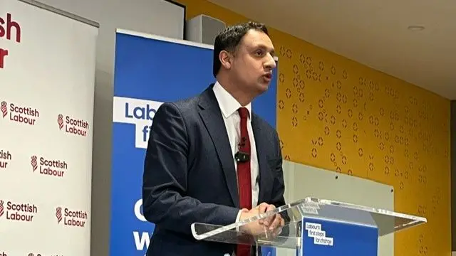 Anas Sarwar speaking with his hands folded on a clear podium with posters saying 'Scottish Labour' and 'Cut NHS waiting times' behind him