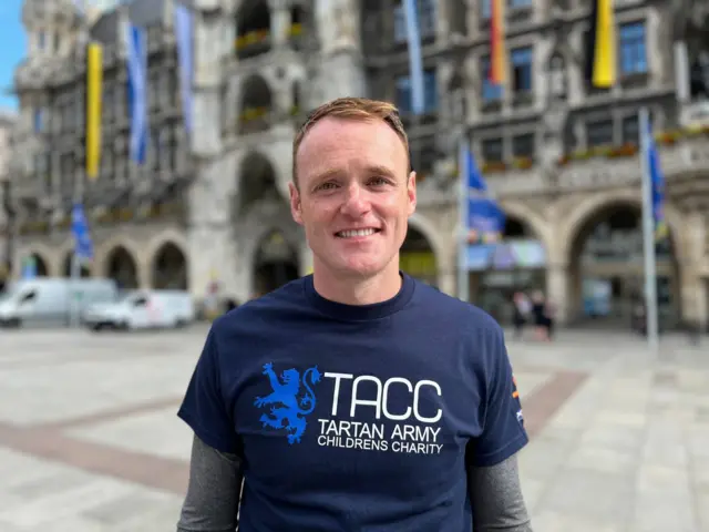 Derek smiles proudly in central Munich with the words 'TACC Tartan Army Childrens Charity' displayed on his blue t-shirt