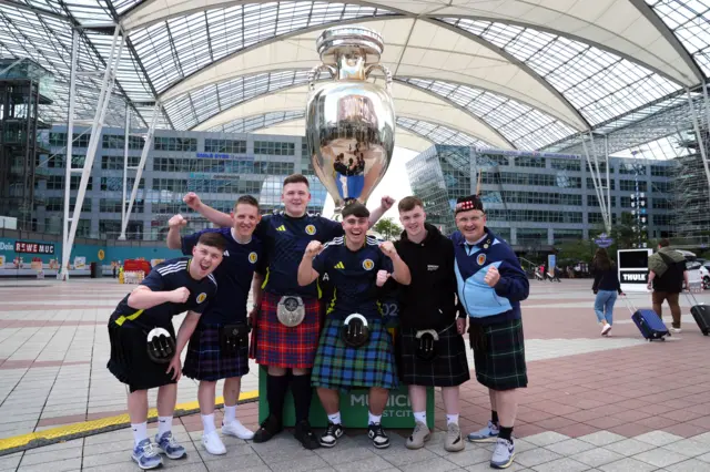 Scotland fans wearing kilts pose in front of a giant European Championship trophy at Munich airport