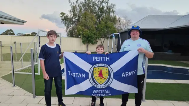 Gary and sons Ross and Greg proudly hold a Scotland flag in a sunny garden with a swimming pool