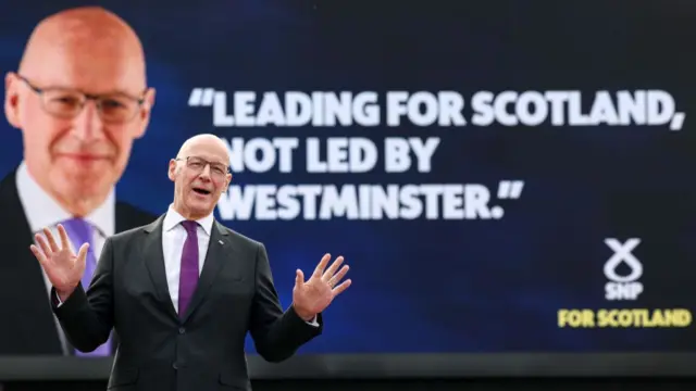 John Swinney in front of a billboard of John Swinney and a quote reading "Leading for Scotland, not led by Westminister"