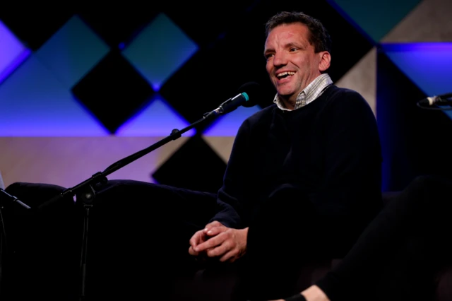 Henning Wehn laughs in front of a microphone at an indoor event