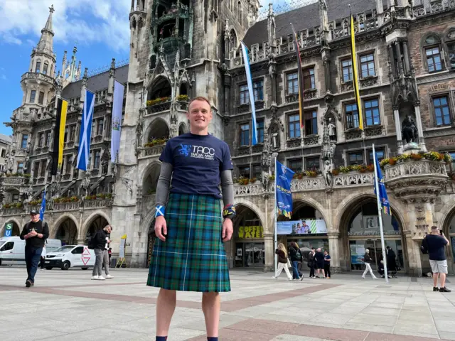 Derek, wearing a kilt, smiles confidently in central Munich with the words 'TACC Tartan Army Childrens Charity' displayed on his blue t-shirt. He also wears a wristband with a Scottish flag.