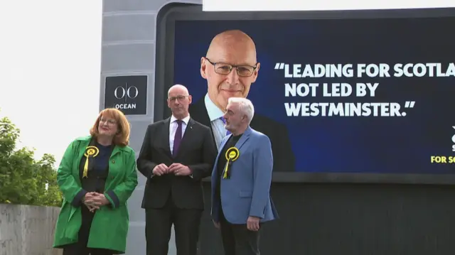 John Swinney standing in front of an SNP poster