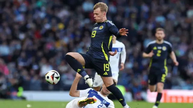 Scotland winger Tommy Conway playing against Finland in a friendly match.  He is skipping past a finland player, who is going in for a slide tackle and Conway is jumping over him with the ball in the air.  He is wearing a navy blue Scotland strip with the number 19 on it.