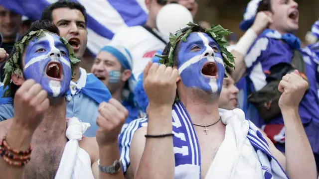 Two greek football supporters with their faces painted in the Greek flag.  They both have their hands high in celebration at a football match at Euro 2024