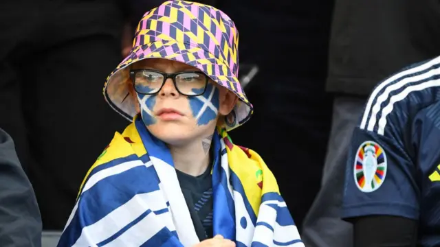 Young Scotland fan wearing a Scotland flag over his shoulders.  He has his face painted with two saltire flags on his cheeks.  He's wearing a pink and yellow bucket hat