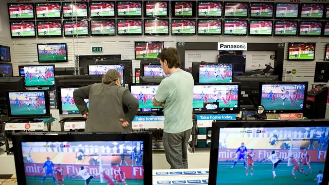 Football fans watching a match surrounded by loads of tv's in a technology ship.  They are both standing with their arms folded watching the game which is displayed across multiple screens