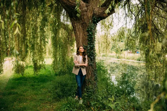 Kate leans against a weeping willow tree dressed in a blazer and jeans