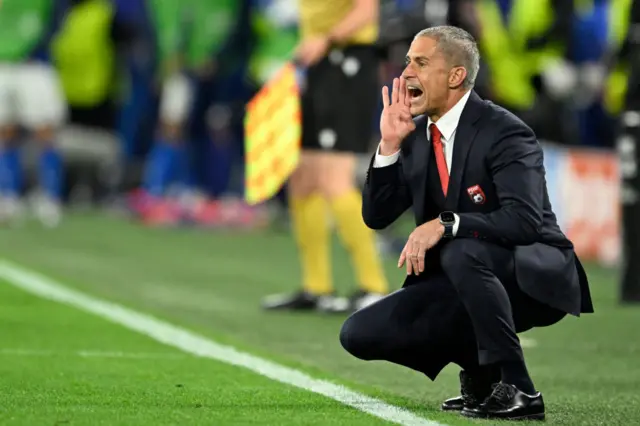 Sylvinho shouts instructions from a crouched position on the touchline