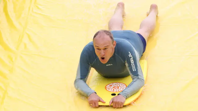 Ed Davey riding on lies prone on a board as he goes down a waterslide