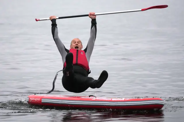 Sir Ed Davey wearing a red life vest, falling off a paddleboard with an oar held in the air