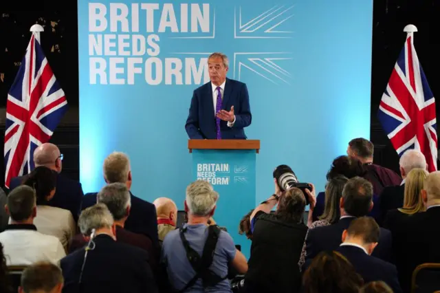 Reform leader Nigel Farage standing on stage behind a lecturn, in front of a light blue sign with the words Britain Needs Reform and flanked by two Union Jacks. He is speaking to a group of people in the foreground