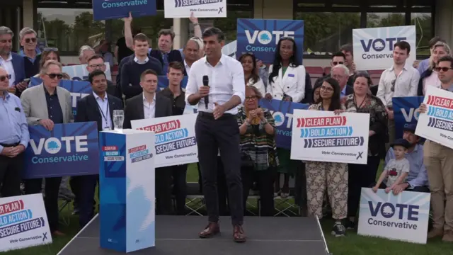 Rishi Sunak stands on a stage in front of people holding signs saying Vote Conservative