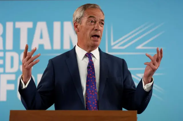 Reform's Nigel Farage standing in front of a light blue Britain Needs Reform sign with his hands animated as he speaks