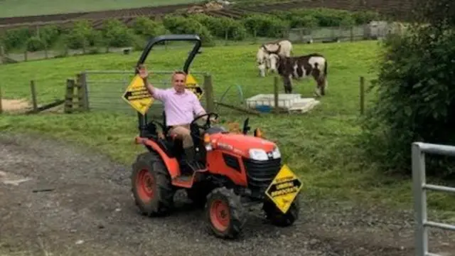 Alex Cole-Hamilton waves as he rides a tractor with Liberal Democrats signage on a farm with animals in the background