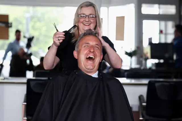 Alex Cole-Hamilton wearing a black smock sitting in a chair, having his haircut as he laughs