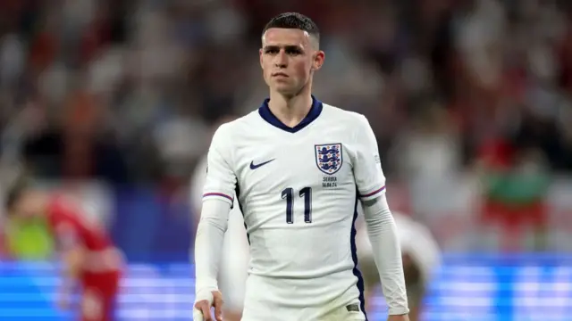 England midfielder Phil Foden wearing a white England strip which has the number 11 on the front.  He has a neutral look on his face