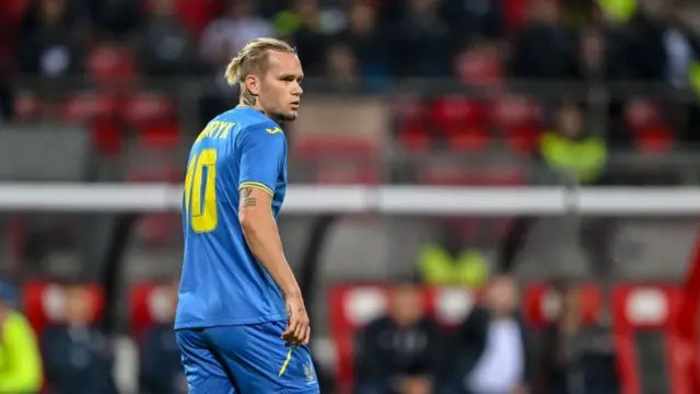 Ukraine strike Mudryk during an international game wearing a blue strip with yellow trimming.  He has a focused look on his face,